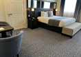 Image shows an accessible bedroom at The Grand Hotel Birmingham. 
A bed with white linen is dressed with a grey blanket and at the foot of the bed is an ottoman in a black and white pattern that closely matches the that of the carpet.  There is a lot of turning space on one side aid the bed and very little room if any for a wheelchair on the other as currently configured.