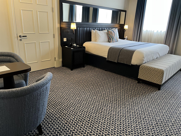 Image shows an accessible bedroom at The Grand Hotel Birmingham. 
A bed with white linen is dressed with a grey blanket and at the foot of the bed is an ottoman in a black and white pattern that closely matches the that of the carpet.  There is a lot of turning space on one side aid the bed and very little room if any for a wheelchair on the other as currently configured.