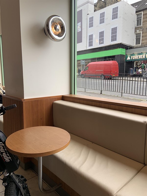 Image of a cushioned booth-style bench with a low backrest with a large window behind it. There is a pillar with a light fixture on one side of it. There is a small circular table that is supported by an angled metal pipe coming from below the seating.