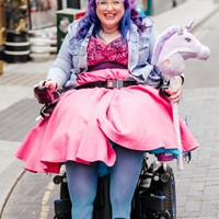 Vikki wears their pink and purple wedding dress sitting in their powerchair. They carry a hobby horse unicorn.