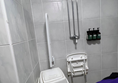 A photo of the shower. A bin is in the shower area. There is only one drop down grab rail. The seat is under the shower controls making both unusable. The overhead shower is out of view.