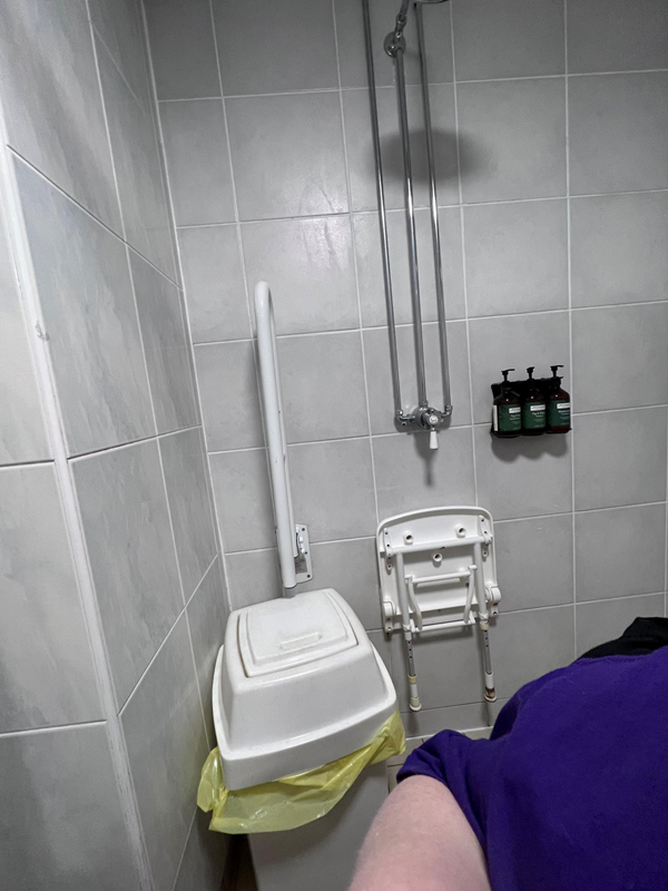 A photo of the shower. A bin is in the shower area. There is only one drop down grab rail. The seat is under the shower controls making both unusable. The overhead shower is out of view.