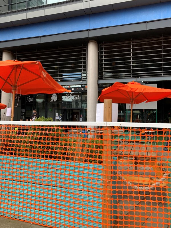 View from outside beer garden. There is orange netting around the plants to stop people walking in between. In the distance a member of staff is wearing a mask.