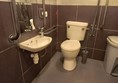 a photo of the bathroom. a sink and mirror are mounted on the wall. to the right of them, a red cord that dangles around a foot off the ground and a toilet with grab bars. a sanitart bin is in the corner.
