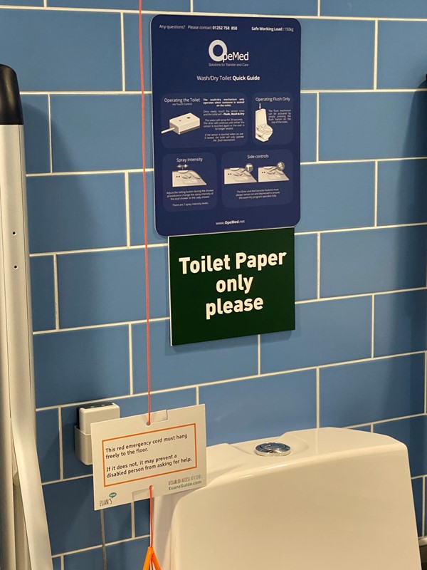 Instructions over the toilet for using the hoist and the wash and dry bidet functions, also a sign saying "toilet paper only please", and a euan's guide red cord card on the red cord which now hangs freely next to the toilet