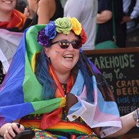 Vikki is smiling, they are dressed in rainbows with a flag around them for a pride parade.