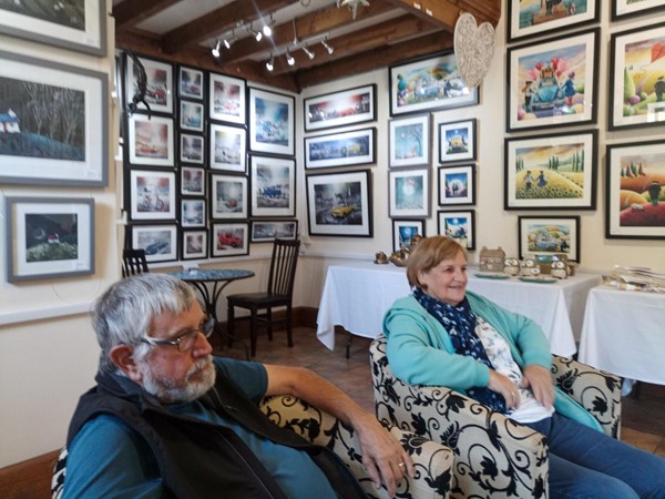 Elderly couple sitting in armchairs, with framed pictures on the walls behind them.