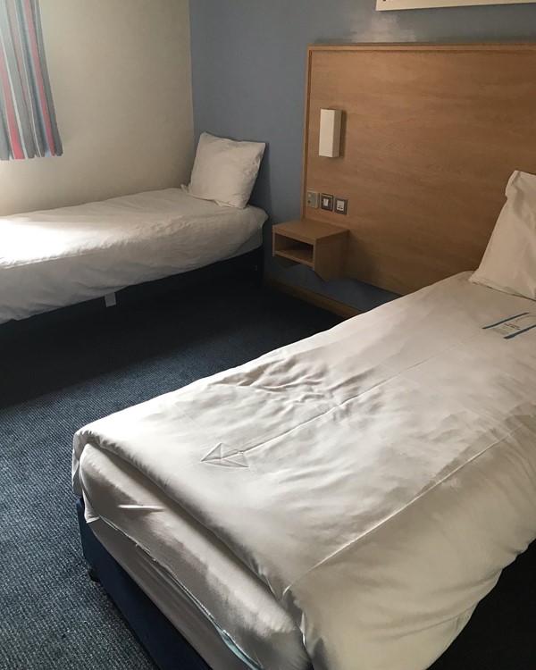 two single beds in hotel room.
one pushed up against a wall with a high window, the other pushed close to a wall.