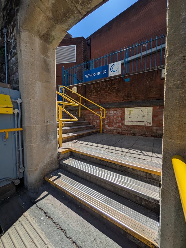 Image of steps leading out of the station, there are three steps, a level platform, then seven more. There are rails on either side, tactile paving and yellow markings at the edge of the bottom and top steps