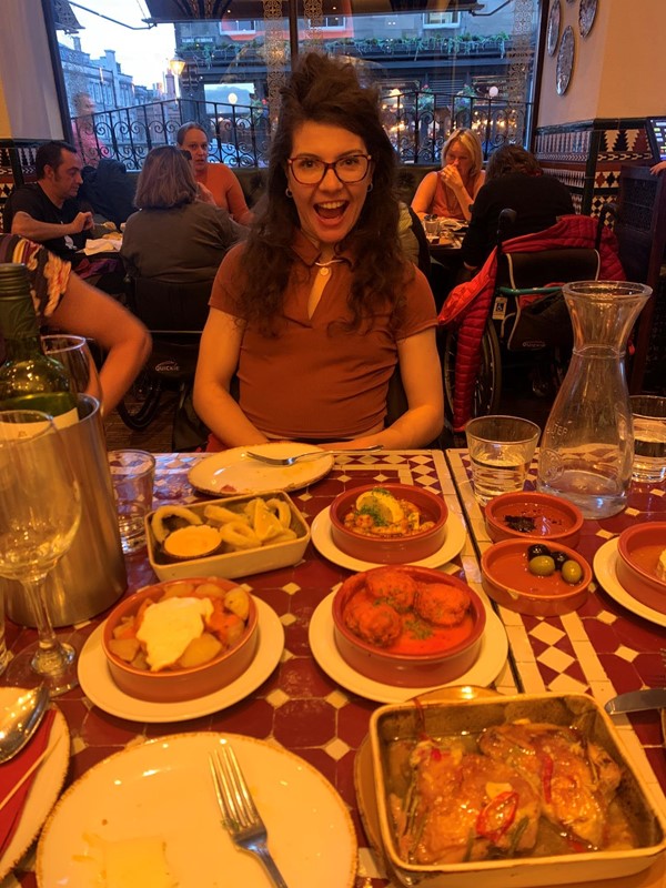 Photograph shows a young wheelchair user at a table with lots of tasty Tapas in front of her. Behind her is another table with a number of wheelchair users sat comfortably