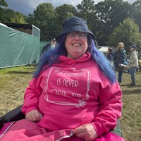 Vikki sits in their powerchair in a festival field. They have a bucket hat and a pink hoodie. The hoodie says ‘Accessibility is never optional’.