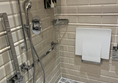 Image shows a wetroom-style bathroom in an accessible room bedroom at The Grand Hotel. There is fairly good colour contrast between the the floor and wall tiles, a chrome shower hose that can be handheld or fixed at a desired height.
There is a white plastic flip down seat and some
amenities fixed to the wall.
