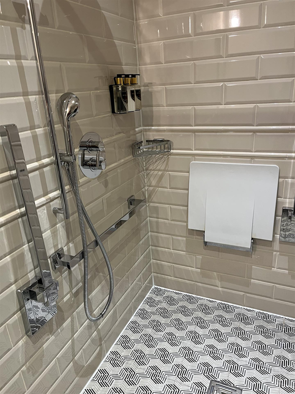 Image shows a wetroom-style bathroom in an accessible room bedroom at The Grand Hotel. There is fairly good colour contrast between the the floor and wall tiles, a chrome shower hose that can be handheld or fixed at a desired height.
There is a white plastic flip down seat and some
amenities fixed to the wall.