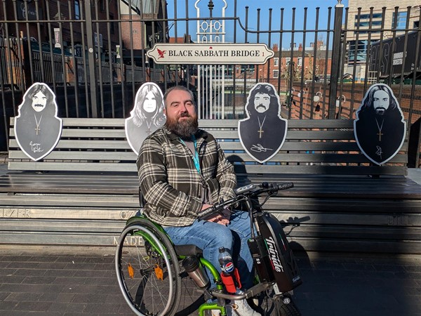 Image of Joe, a wheelchair user, in front of Black Sabbath Bridge, which shows  all band members and their signatures.