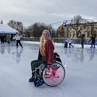 Roz is ice skating in their hospital chair, which has pink spoke covers on the wheels. they are wearing ice skates and are smiling at the camera. the ice rink is in Greenwich and the historic buildings of the Old Royal Navel College are visible.