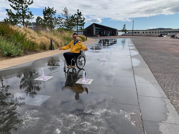 Paul sits in his wheelchair in the middle of the paved area with jumping fountains squirting up from the paving. His is smiling and has both hands giving a thumbs up