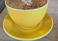 Image of a large cappuccino in a yellow cup with a matching saucer beneath. Both rest on a light wood table.