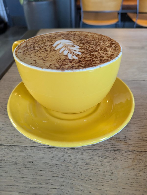 Image of a large cappuccino in a yellow cup with a matching saucer beneath. Both rest on a light wood table.