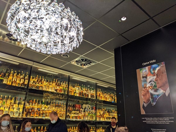 A chandelier with loads of bottles of whisky in the background in a cabinet and a picture of Claive Vidiz drinking whisky to the right.