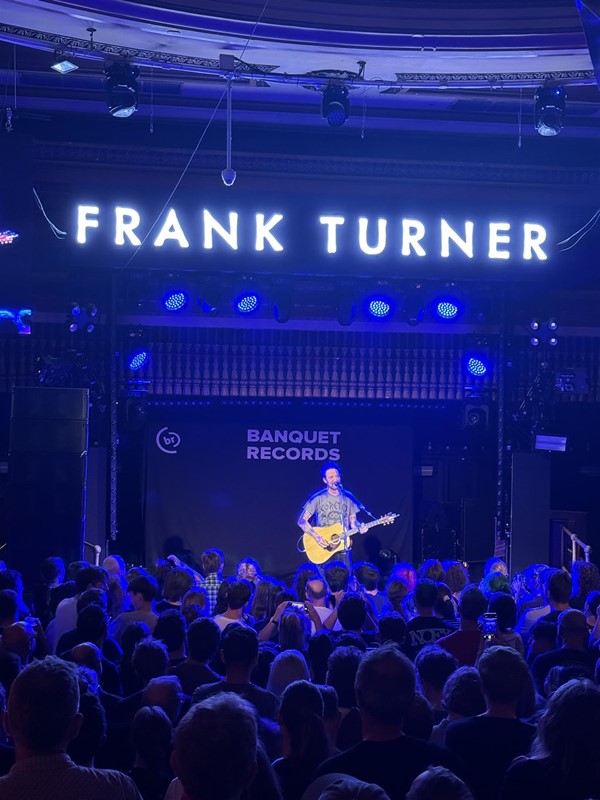 View of artist (Frank Turner) on stage. His name is lit up above him and the crowd standing can be seen.