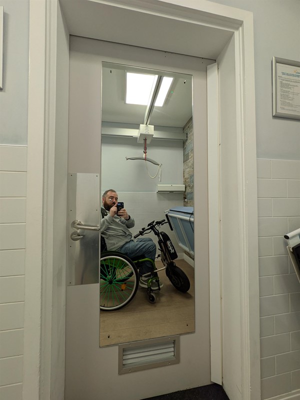 Image of door to the Changing Places toilet from the inside, a white man with dark hair, a grey jumper and blue jeans is reflected in the mirror on the back of the door