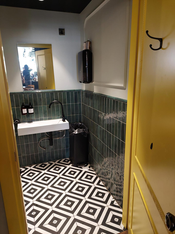 A view of the accessible toilet, looking through the open door, the easy-to-use sink is visible, as is the beautiful geometric-tiled floor. The mirror is standing height and the hand towlels are within reach but above the sink.