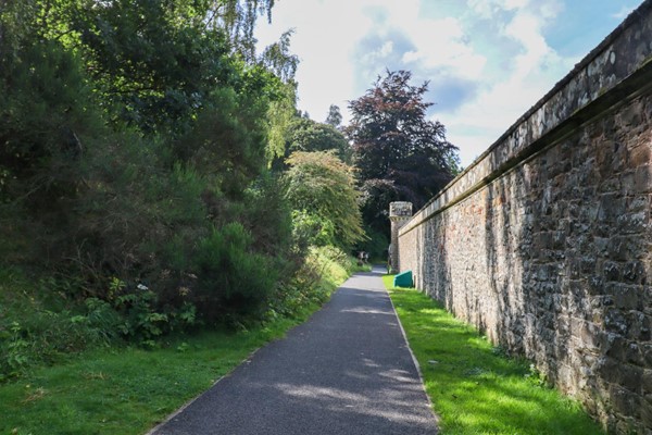 Path from visitor centre to house with a high wall on one side and various bushes on the other.