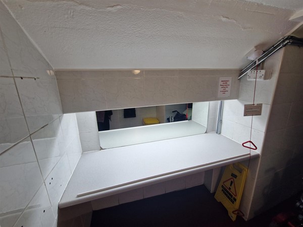 View of the changing table in the accessible toilet. This section would not be locked. A mirror is mounted above it, and the space is brightly lit. A red emergency cord with a Euan’s Guide red cord card hangs nearby.