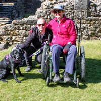 Lady in mountain trike with man and assistance dog in front of abbey ruins.