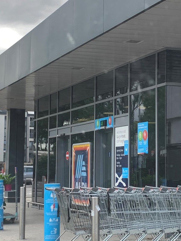 Exterior of shop. The green/red light is above the entrance door. Shopping trolleys are on the right hand side of the door.