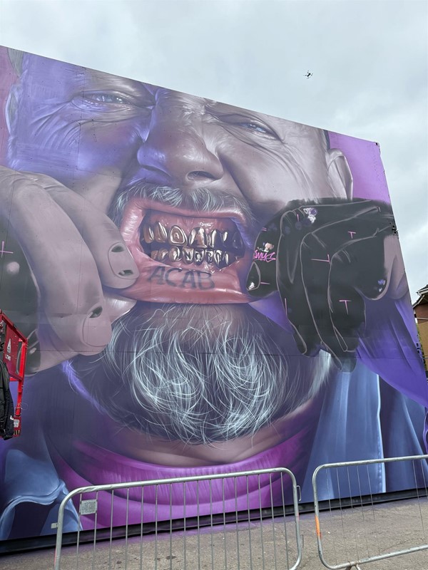 This is a MASSIVE artwork of a man's face with grills on his teeth. He looks like he is crying and he is opening his mouth with his hands to show the grills and the word ACAB on his inner bottom lip.. He has a grey beard and moustache. You can just see the collar of his purple t shirt.