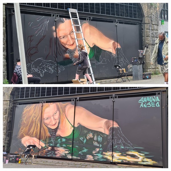 These are 2 photos of the progression of an artwork on boards. It's of a lady with red hair lying down next to a pool of water. She has a green slip dress on. She has her hands gently in the water looking for her ting. She is looking down into the water and looks pensive.