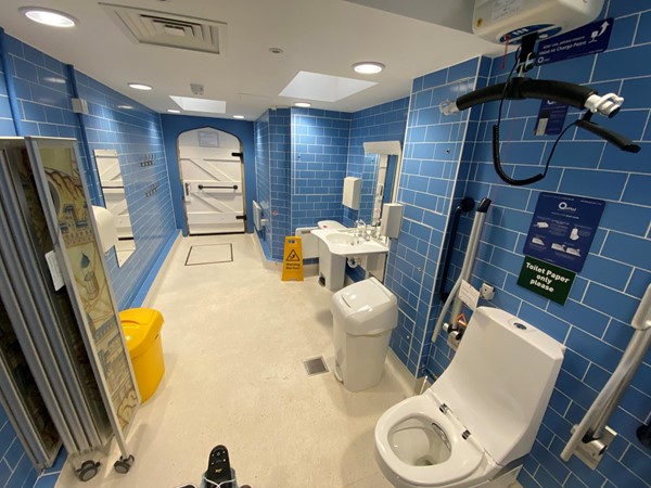 A view of the bathroom taken from the changing/washing table, looking toward the door, showing that it is built in an old building, with an ancient tudor style wooden door, but entirely modern and tiled floor to ceiling - it is impeccably clean with all modern ameneties and looks brand new. There are no windows in the walls due to the location sandwiched between two buildings, but there are a couple of roof light windows in the ceiling letting in ample light even if the electric lights were not to work. The folding screen has a historic scene printed on it, which is a nice touch and something interesting to look at while you're in there. The toilet has a hoist above it, there are at least three different bins in the room, and various grab handles,