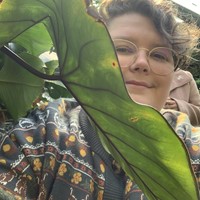 The photo shows Pat, a white person with short brown hair wearing a cardigan with a nature pattern. They are looking out from behold a large green leaf that is growing at chest height.