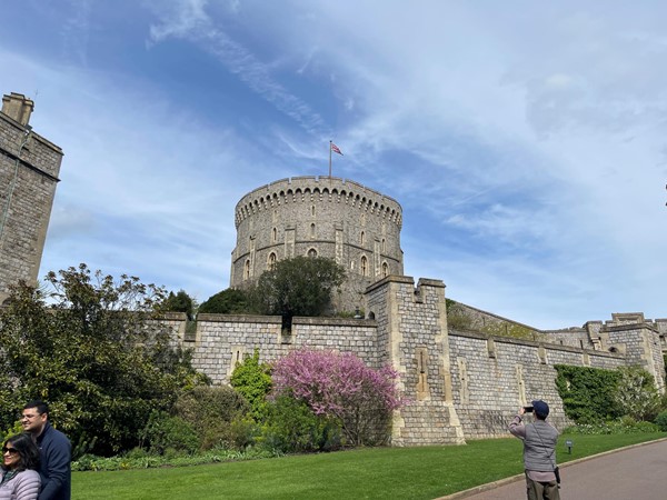 outdoor view of the large bailey tower in the centre of Windsor castle, built from stone constructed on a tall hill. The sky is blue with wispy clouds and there are colourful well-tended plants in bloom, a lovely spring day, some other tourists are walking through the shot. The path is smooth.