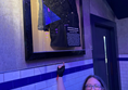 A white woman with long brown hair and glasses points up at a jacket worn by Stars from Bang Bang Romeo. She wears a dress with dinosaurs on and a glove on her pointing hand.
