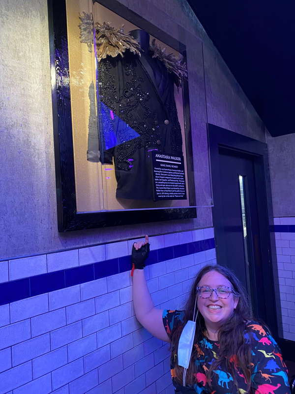 A white woman with long brown hair and glasses points up at a jacket worn by Stars from Bang Bang Romeo. She wears a dress with dinosaurs on and a glove on her pointing hand.