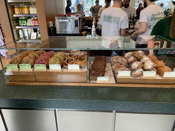 Image of a glass display case on top of a counter containing baked goods. They are (from left to right): Carrot Cake Cookie, Sea Salt Milk Chocolate Cookie, Raspberry White Chocolate Cookie, Pistachio Creme Cookie, Cinnamon Bun, Chocolate Banana Bread (vegan), Morning Bun, Almond Croissant, Croissant. The signs are written in all upper-case letters and also show the prince and calories. There is a fridge with Lemonaid and water to the left in the background.