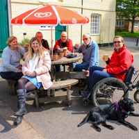 Multigenerational family sitting round an outdoor coffee table with parasol. One of the group is using a mountain trike and has an assistance dog lying in front of her.