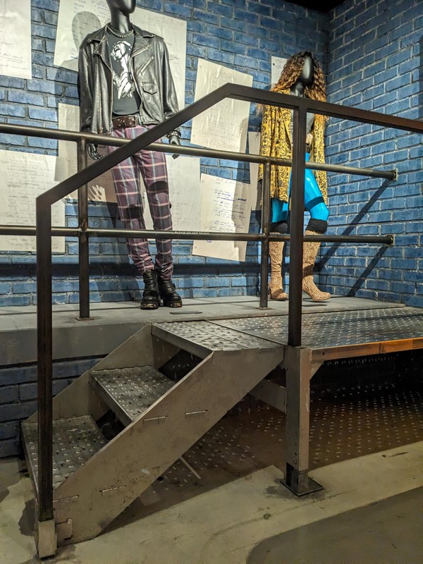 Katie is trying to look in a mirror that belonged to Jonathan Larson but the MOB have decided to set it atop a staircase for little apparent reason.