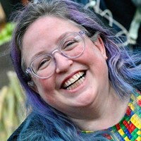 A white person with long purple and blue hair is laughing. They wear glasses and a Lego design top.