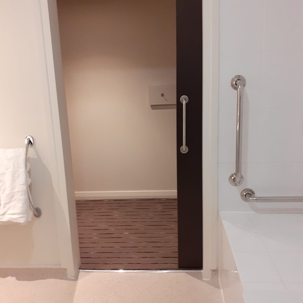The sliding door to the bathroom is open looking out to a beige wall. There is a lowered bath on the right with a place to sit and bars to hold on to.