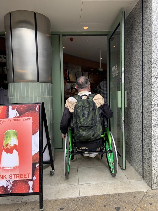 Image of a wheelchair user entering into the coffee shop through the door on the right. The door is not much wider than the wheelchair.