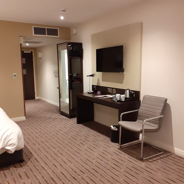 An accessable hotel room with a double bed on the left. Along the right wall is a chair, a table with wall socket, kettle, mugs, and a lamp. There is a TV above the table and a wardrobe with a mirror on the front too.