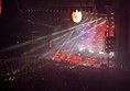Ovo stage with red lighting and search lights going out over the heads of a full standing area of fans.

Fall Out Boy are on the stage plating instruments.