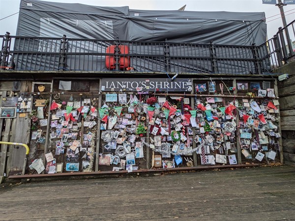 Image of the shrine, white letters on a black sign read "Ianto's Shrine" and there are plenty of pictures and tributes.