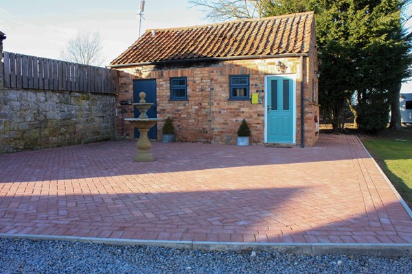 Ladies toilet with a small step and narrow door, which is closed. The area in front is large and paved, with a wide path going round the back of the building to the elsan point.