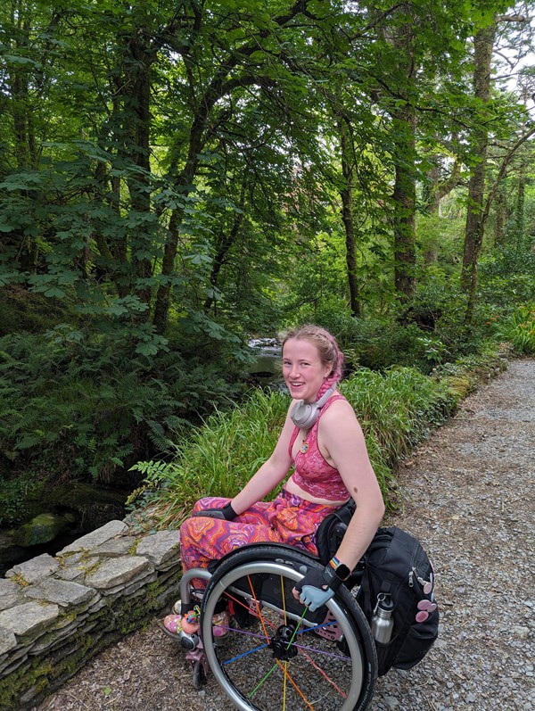 A photo of me enjoying the gardens!

Roz (a white person with pink hair, wearing a pink top and patterned trousers) sits in their manual wheelchair next to a river. Trees are in the background. They are on a gravel path.
