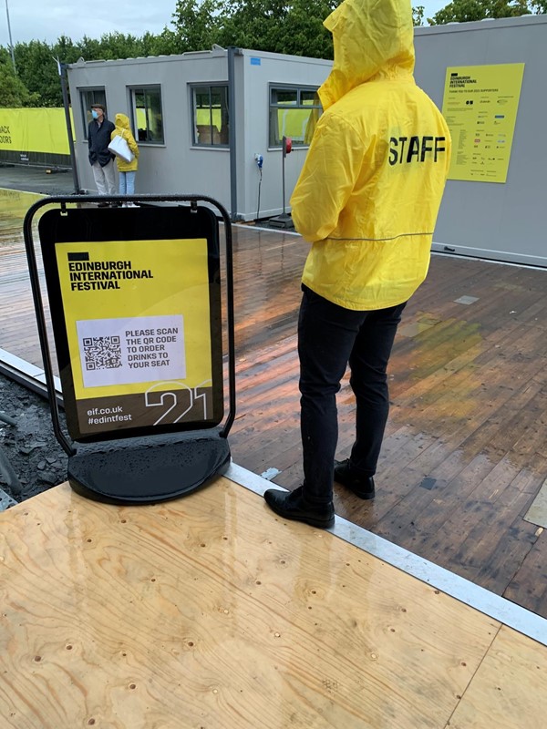 Member of staff standing by a sign saying please scan QR code to order drinks from your seat