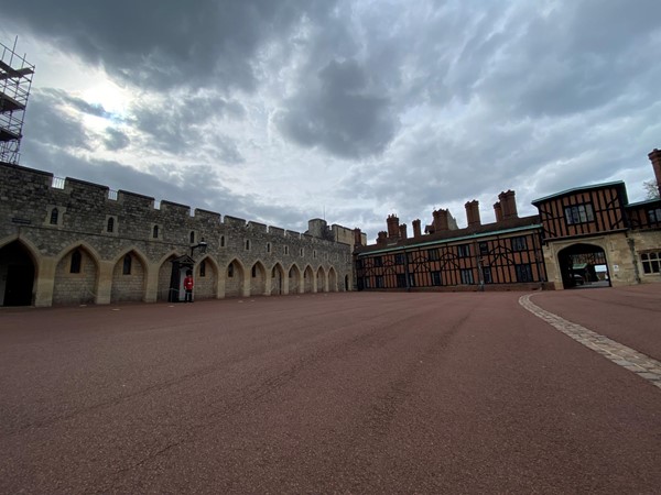 outdoor photo showing an area of the castle with a large smoothly paved courtyard, a traditional guard with red coat and black bearskin hat stands on sentry by a guard box. A half timbered and red plastered tudor building is at right angles to the stone built wall area with blind arcading and small windows with crenellations on top.